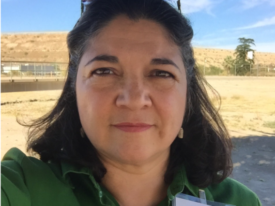 Photo of a person in a green blouse with a printed Project Harvest nametag in front of a field and a fence line in background